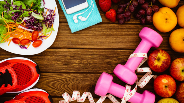 An overhead view of a table with trainers, a plate of salad, headphones connected to a phone, fruit and a pair small dumbbells laid on it
