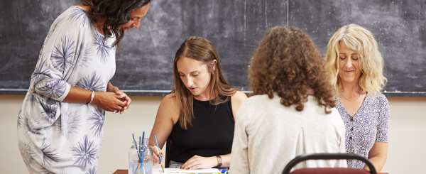 Three women sit around a table taking part in an art class. An instructor stands and looks at their paintings