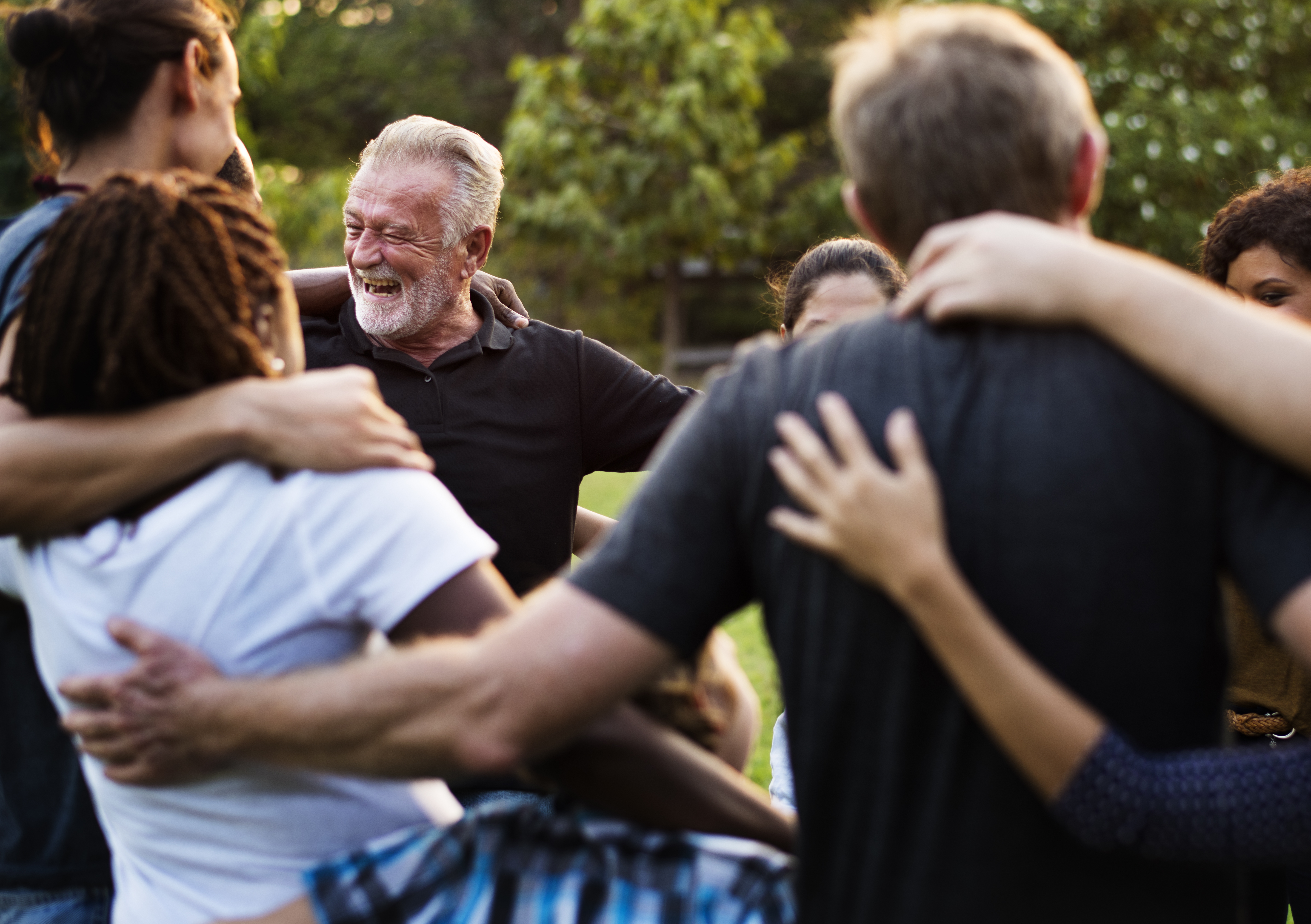 A group of adults of varying ethnicities link arms while standing in a circle at a park