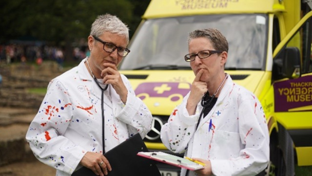 Two women wearing white doctors coats with paint stains pose with their hands to their mouths