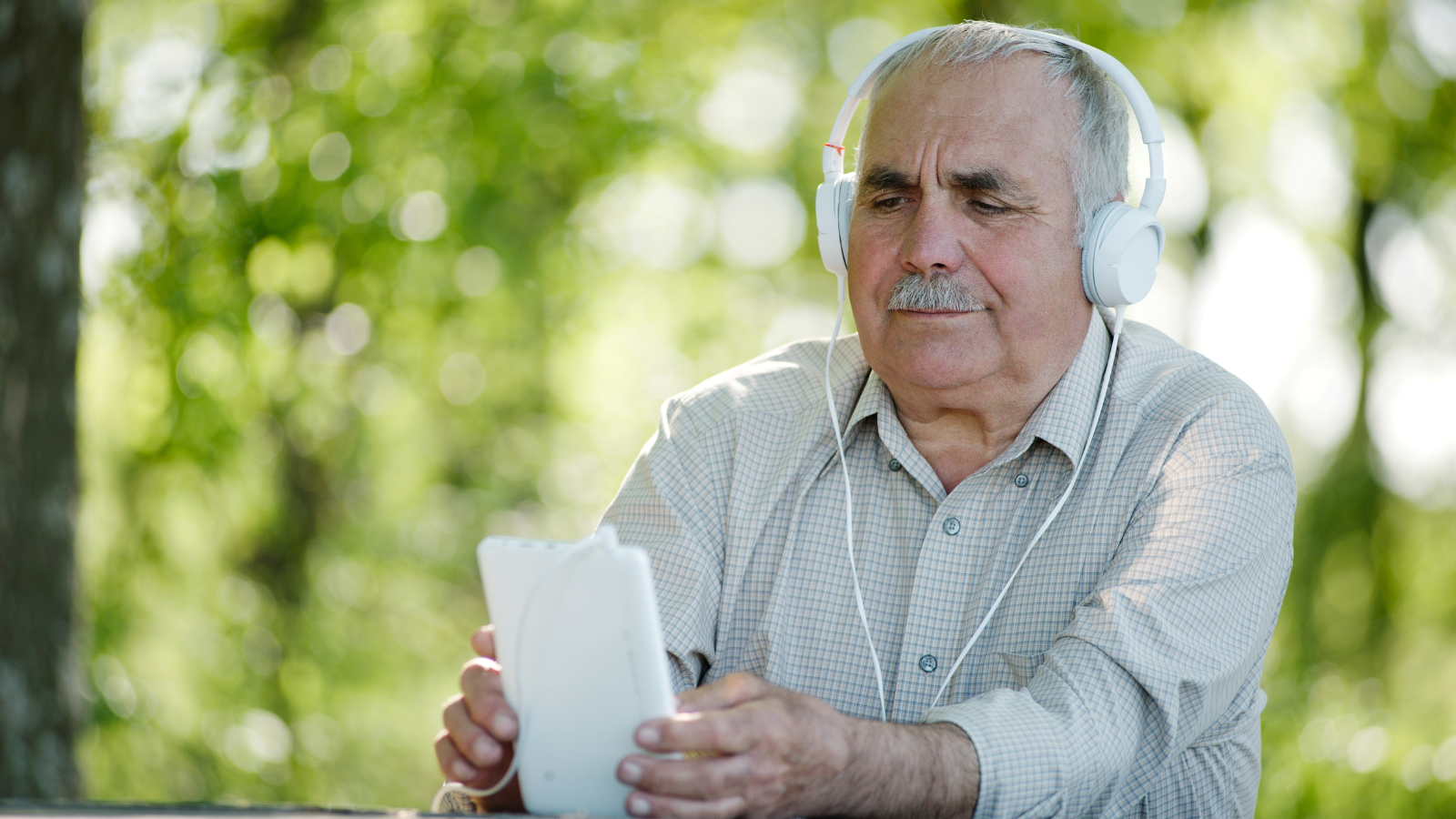 An older man sits at a table with trees in the background. He wears headphones while reading a tablet.