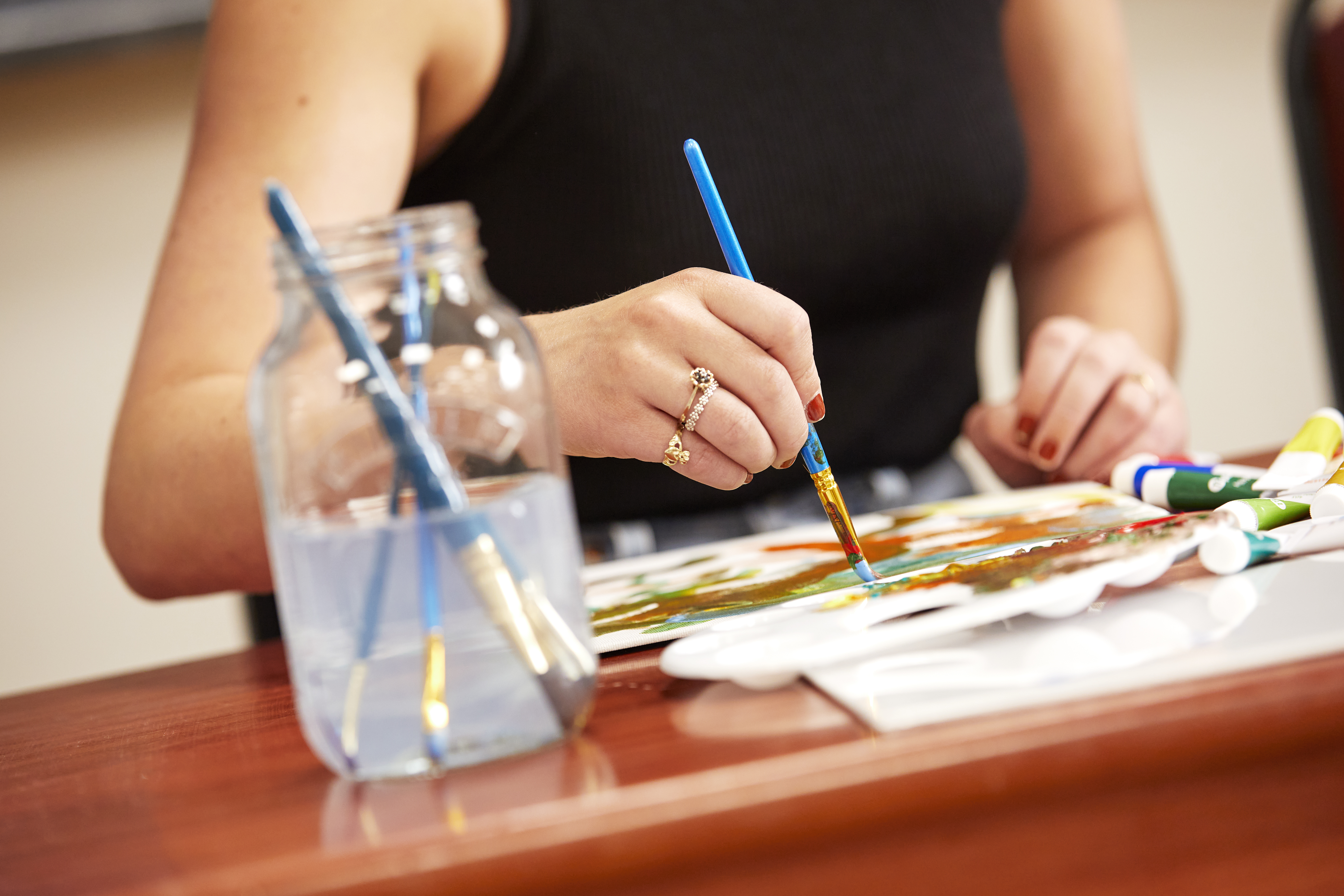 A close up of someone painting on a canvas with a water jar of brushes 