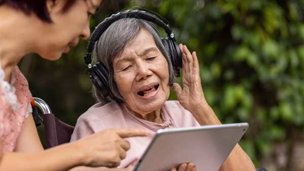An elderly lady in a wheelchair wearing headphones and smiling with her daughter showing her something on a tablet