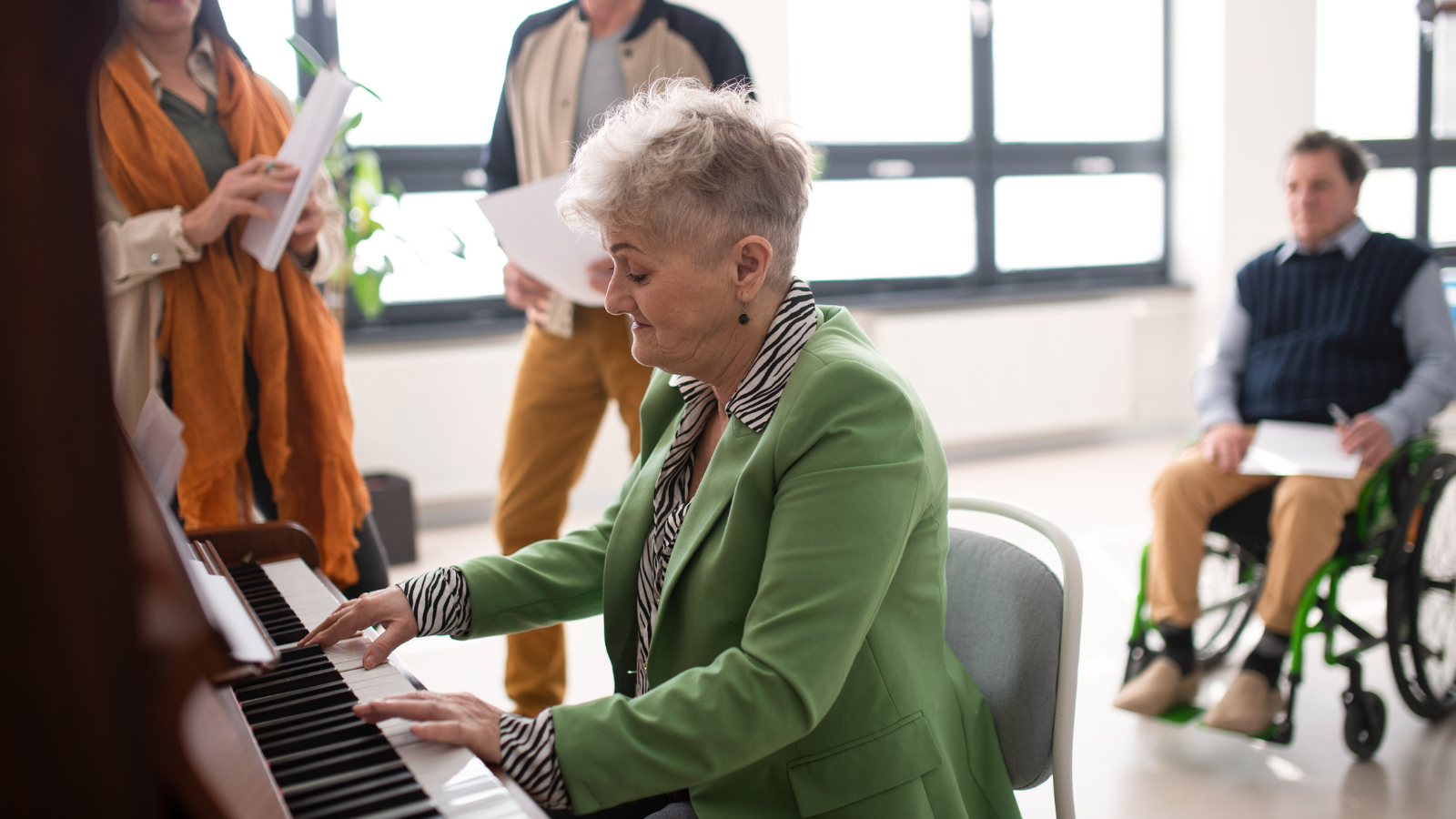 An older lady is sitting at a piano an playing music, behind her a group of friends are reading along to the music and smiling together