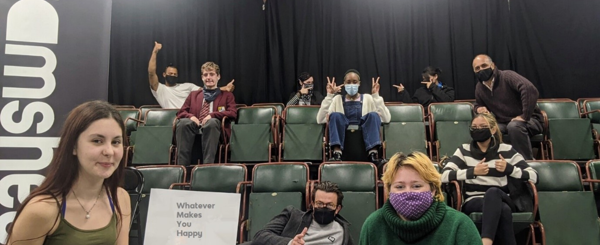 A group of young people sit in the audience at a theatre and hold up a sign that reads 'whatever makes you happy'