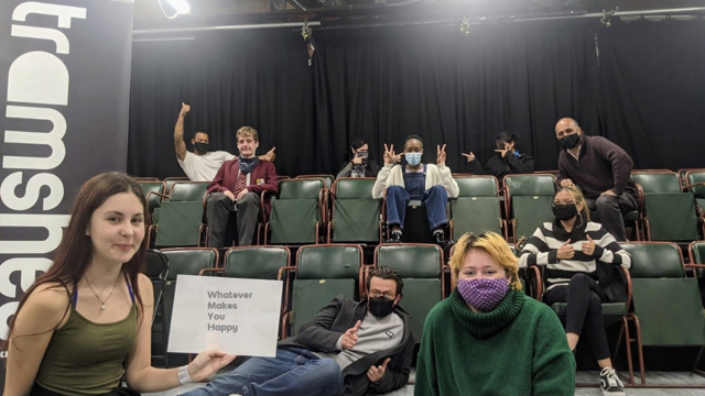 A group of young people sit in the audience at a theatre and hold up a sign that reads 'whatever makes you happy'