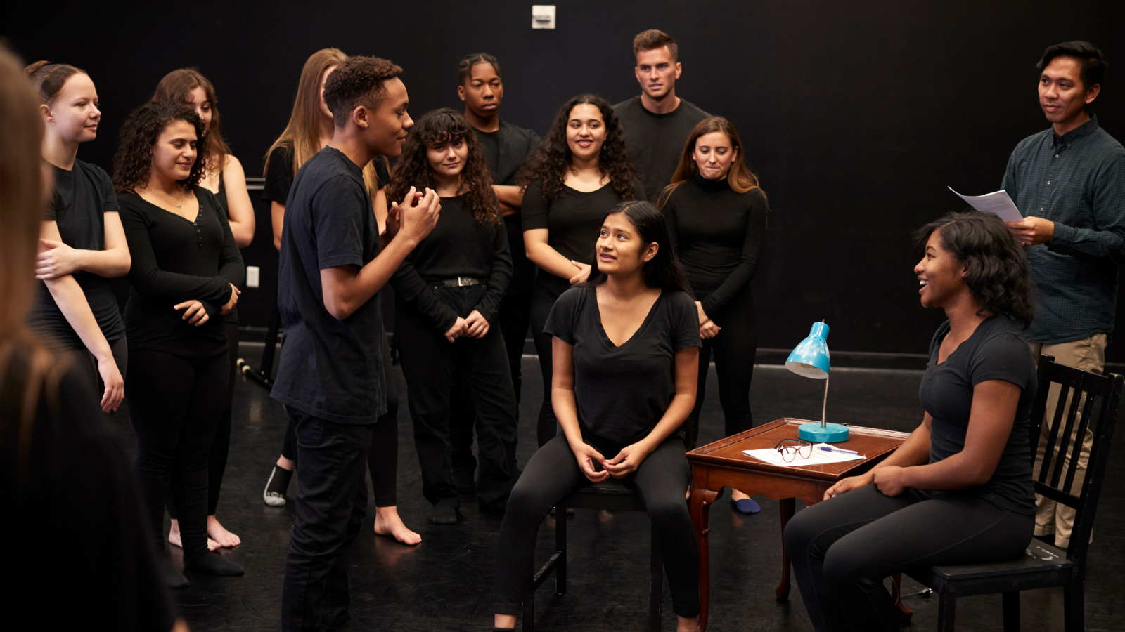 A group of ethnically diverse young people who are dressed in black, rehearse a scene from a play.