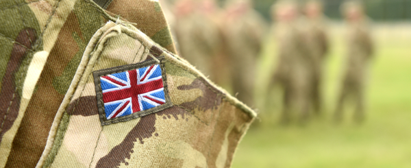 Closeup of a British Soldier's camouflage uninform showing a small Union Jack. A group of soldiers stand in a grassy field in the background.