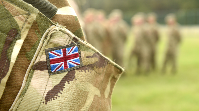 Closeup of a British Soldier's camouflage uninform showing a small Union Jack. A group of soldiers stand in a grassy field in the background.