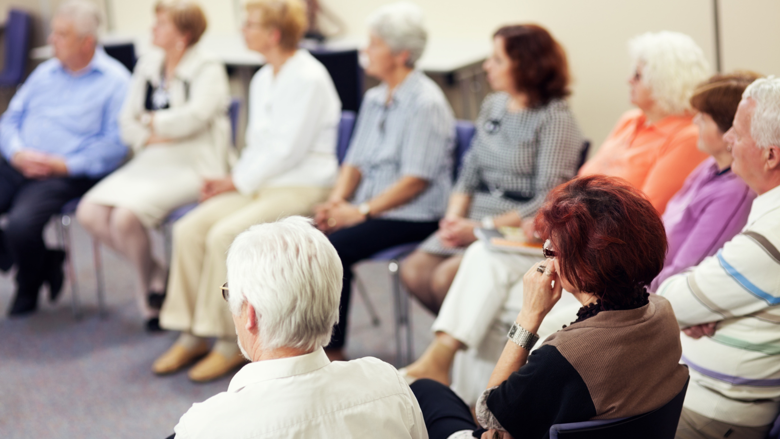 A group of older adults sit on chairs in a circle and listen to a speaker at the community centre