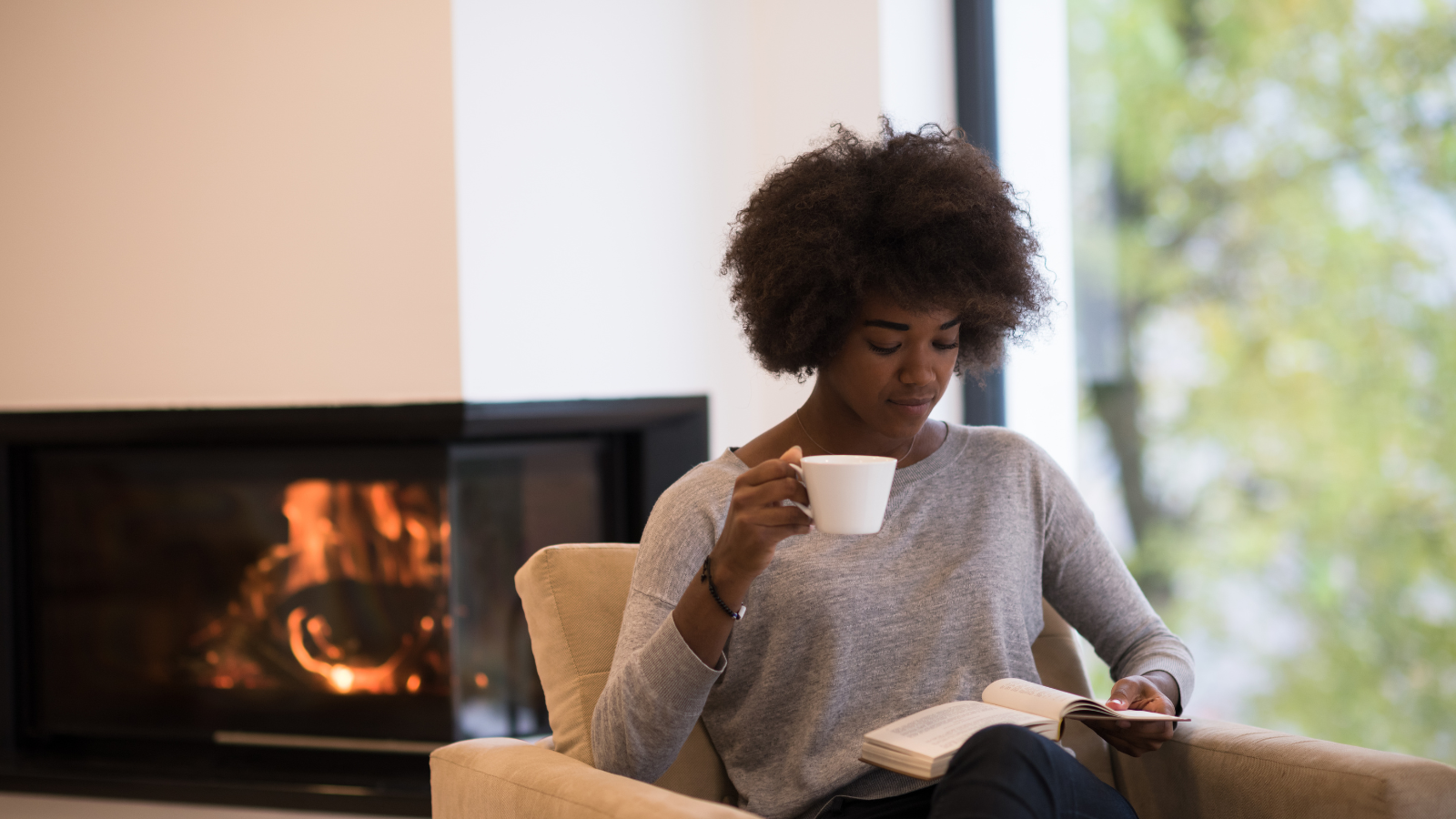 A young woman is sitting in an armchair and is reading a book while holding a mug. A woodfire is in the background