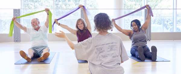 Three people in an exercise studio sit on mats holding resistance bands above their heads, facing an instructor