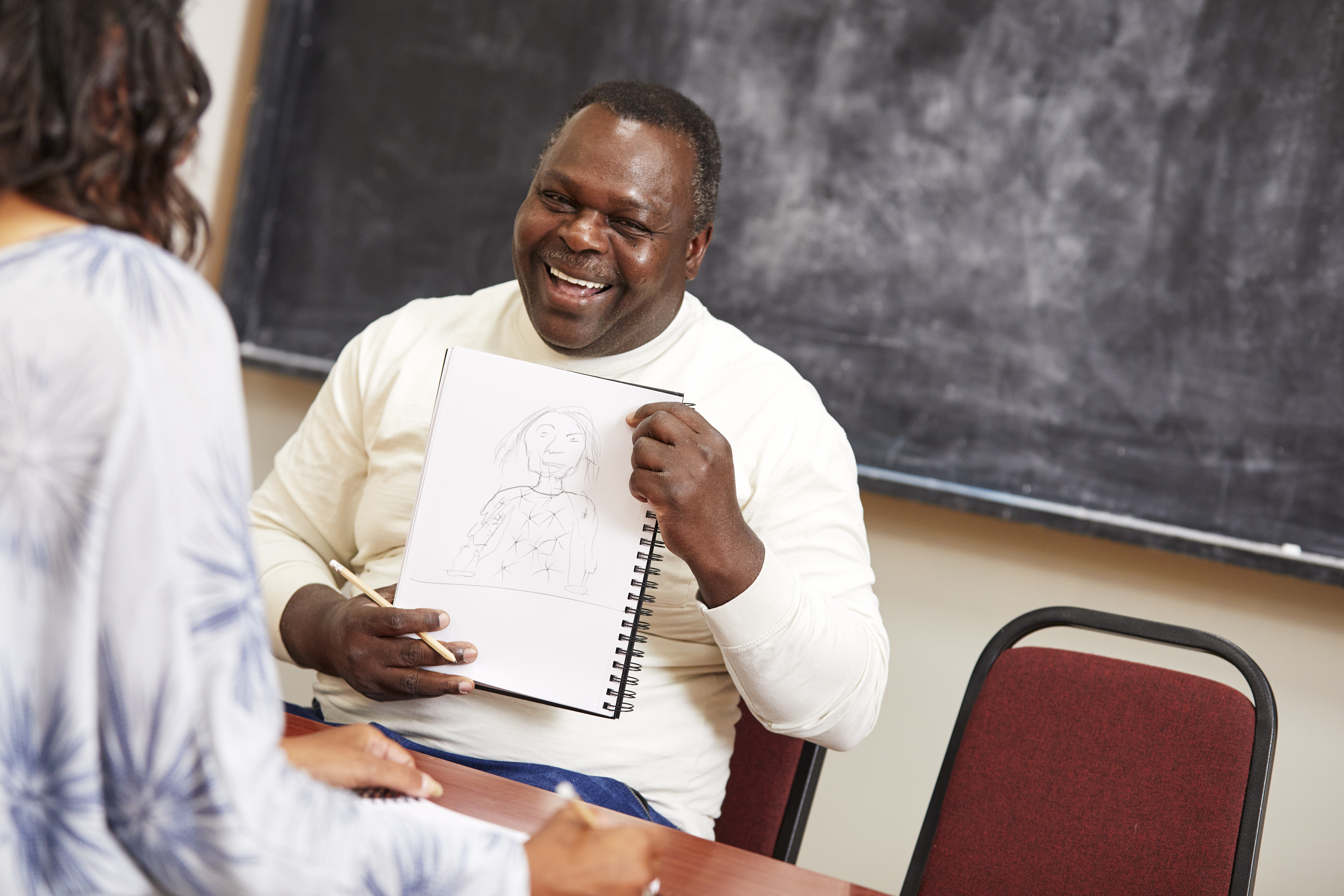 A Black man sits opposite a woman and holds up a notepad, showing her the drawing he has made of her