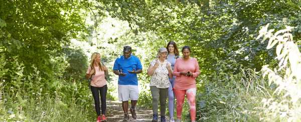 A group of people of varying ages and ethnicities walk and talk together along a sunny wooded path 