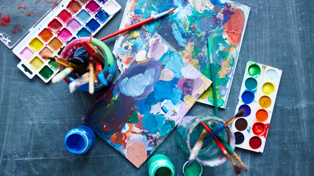 Paintbrushes, pallets and colourful abstract artwork sits on a table