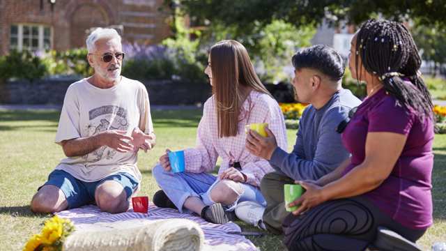 A group of people of varying ages and ethnicities sit on a picnic blanket drinking from mugs.