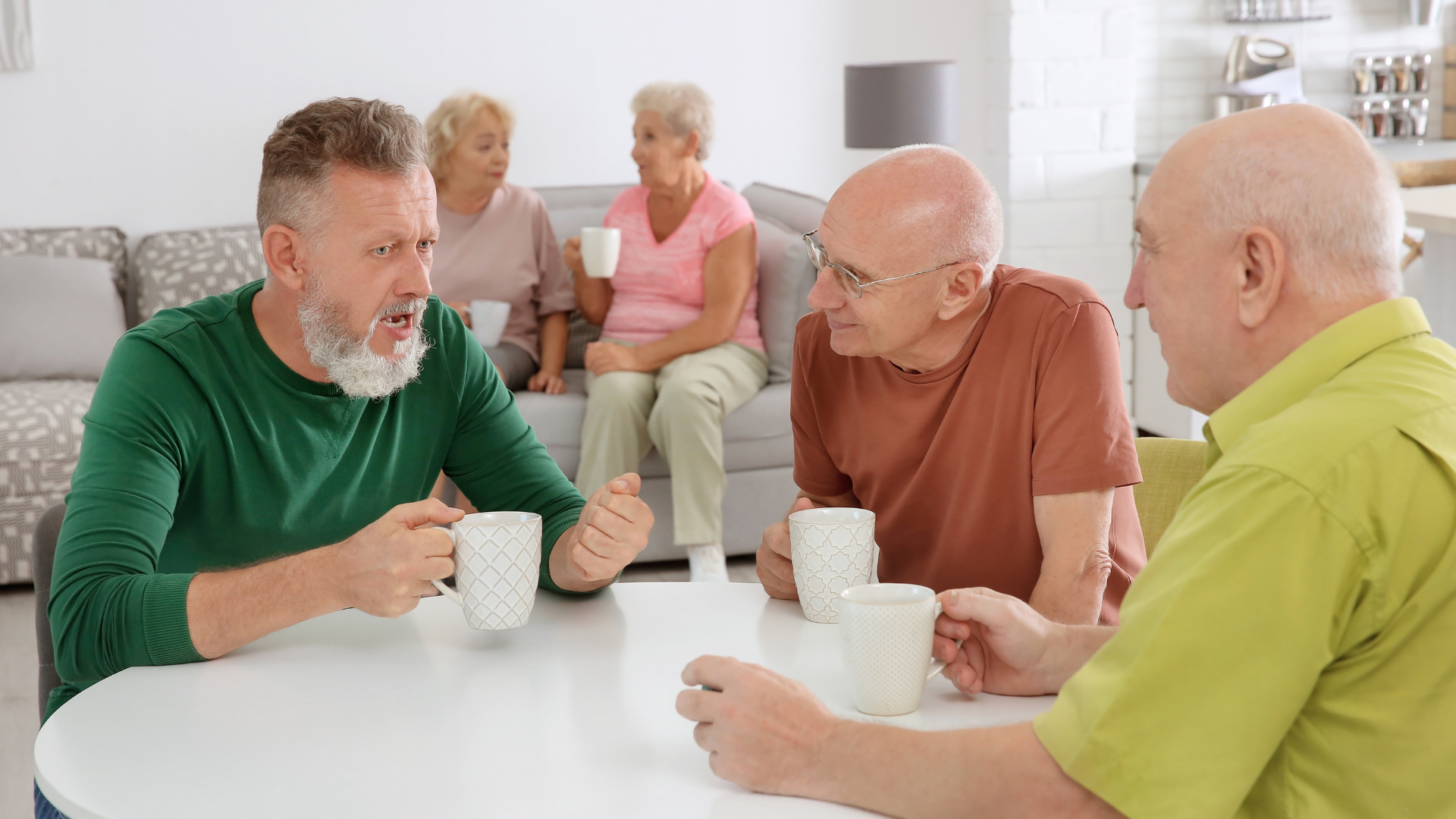 3 older men sit at a table with cups of tea chatting. Two elderly women are in the background chatting on a sofa 