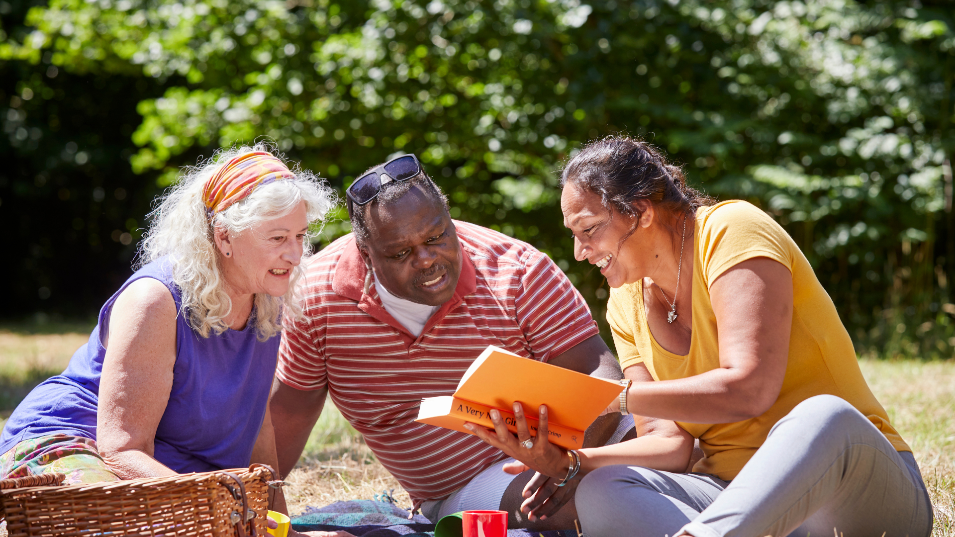 Two women sitting with a man in a park, one woman is reading a book and the others are looking at it as well