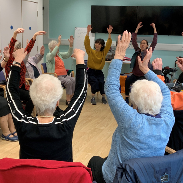 A group of older people sit in a circle with their arms raised during a seated exercise 