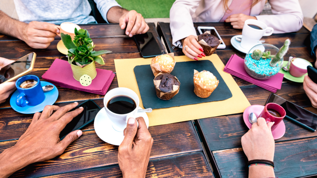 A group of people sit around a table with coffee mugs and muffins. Their phones are visible 